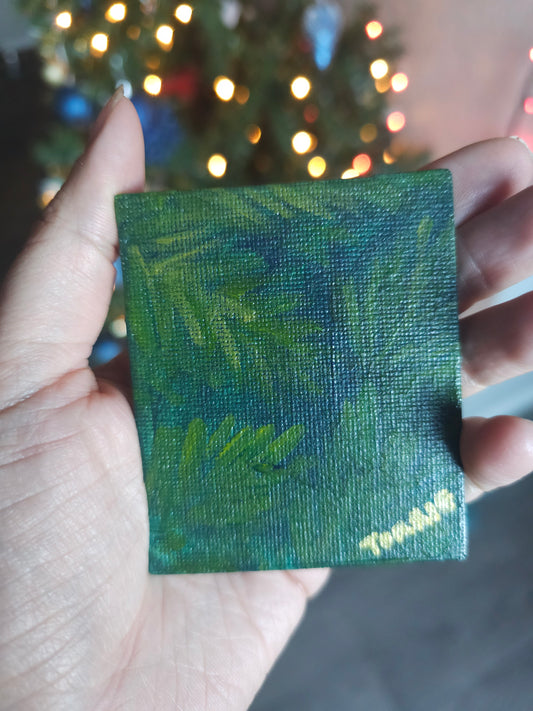 Hand holding a small green textured  tree branch painted on a square magnet with blurred Christmas lights in the background